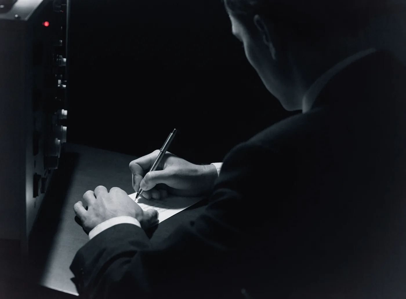 Persona escribiendo a mano en un escritorio en penumbra, junto a un panel técnico apagado con una sola luz roja: la decisión la toma alguien